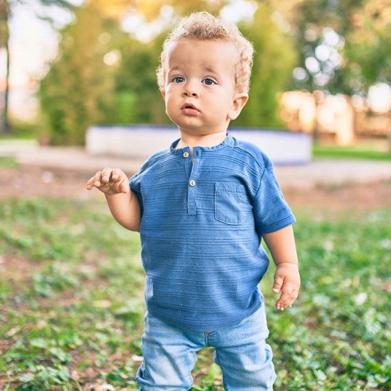 Kleiner Junge in blauer Kleidung steht auf Gras, schaut neugierig.