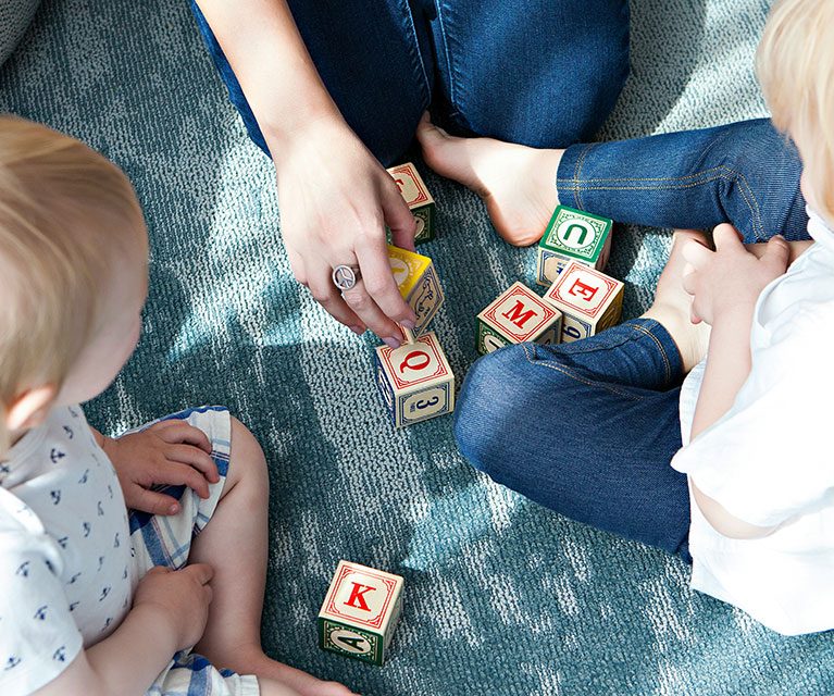 Verschiedene bunte Holzbausteine auf einem Teppich, zwei Kleinkinder spielen damit.